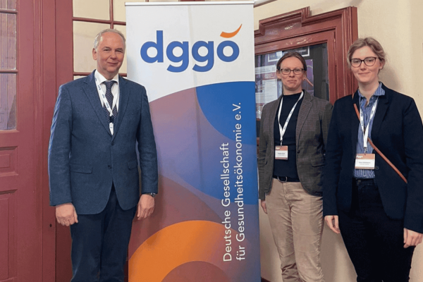 DGGÖ Jahrestagung Gruppenbild (2) (2) Prof. Steffen Fleßa, Dr. Claudia Hübner und Lena Schmeyers stehen neben einem Roll-up-Banner der Deutschen Gesellschaft für Gesundheitsökonomie