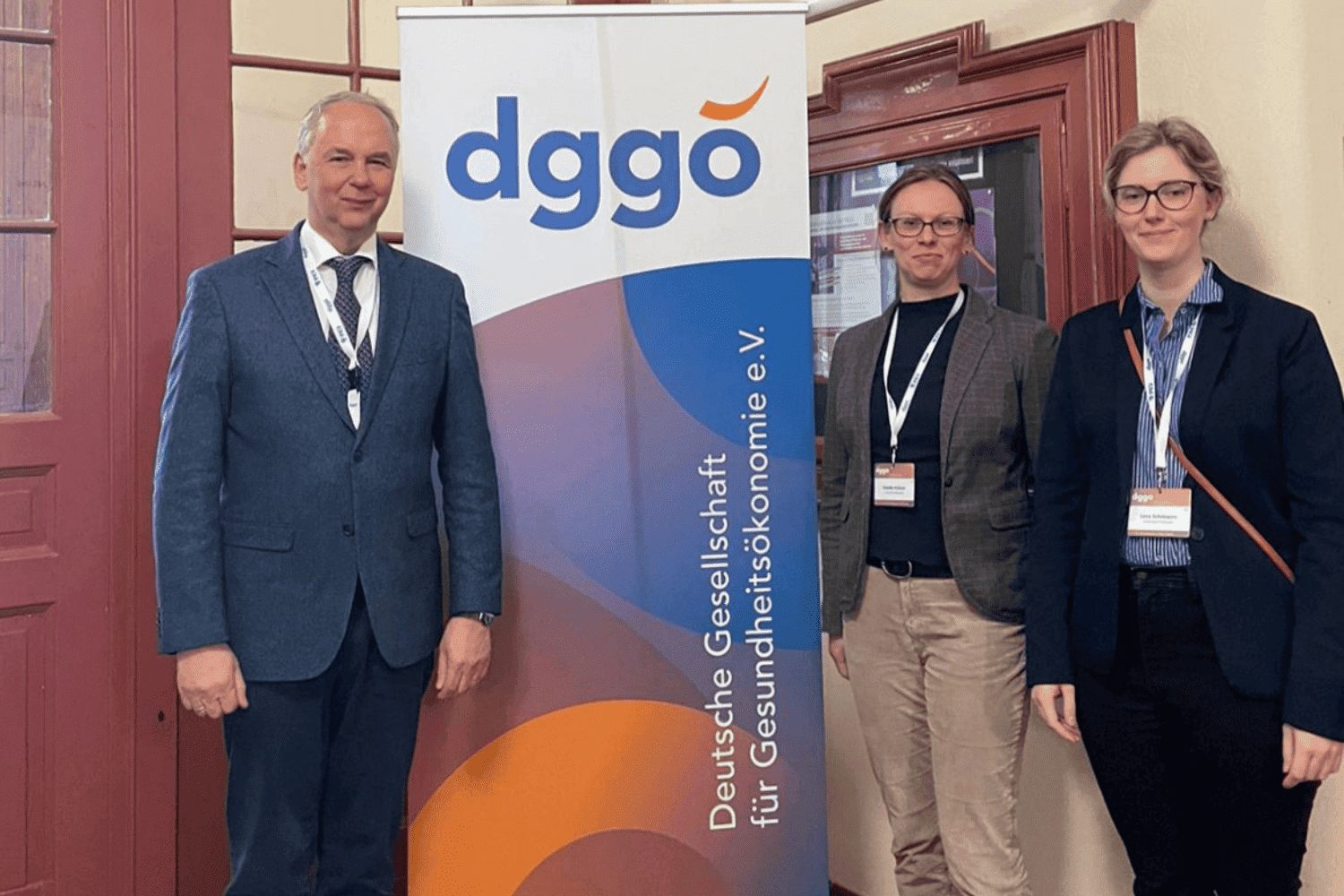 Prof. Steffen Fleßa, Dr. Claudia Hübner und Lena Schmeyers stehen neben einem Roll-up-Banner der Deutschen Gesellschaft für Gesundheitsökonomie