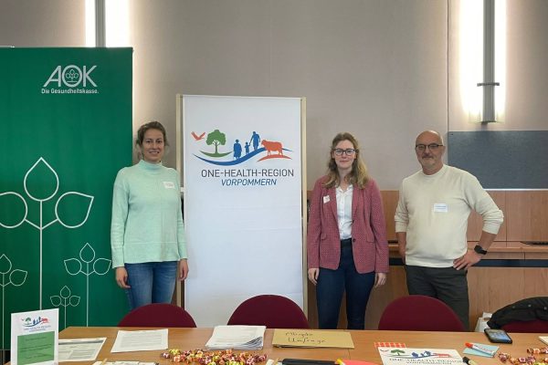 Drei Vertreterinnen und Vertreter der One-Health-Region Vorpommern stehen am Informationsstand beim Markt der Möglichkeiten der 4. Kommunalen Gesundheitskonferenz in Grimmen. Im Hintergrund sind ein Roll-up der One-Health-Region Vorpommern sowie ein Banner der AOK zu sehen.