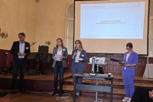 Vier Personen stehen auf einer Bühne und moderieren eine Veranstaltung. Im Hintergrund ist eine Präsentationsfolie mit dem Titel „Zwischen Idee und Realität: Gesundheitsförderung vor Ort starten!“ zu sehen. Die Szene zeigt eine Podiumssituation in einem historischen Saal mit Technik und Leinwand.