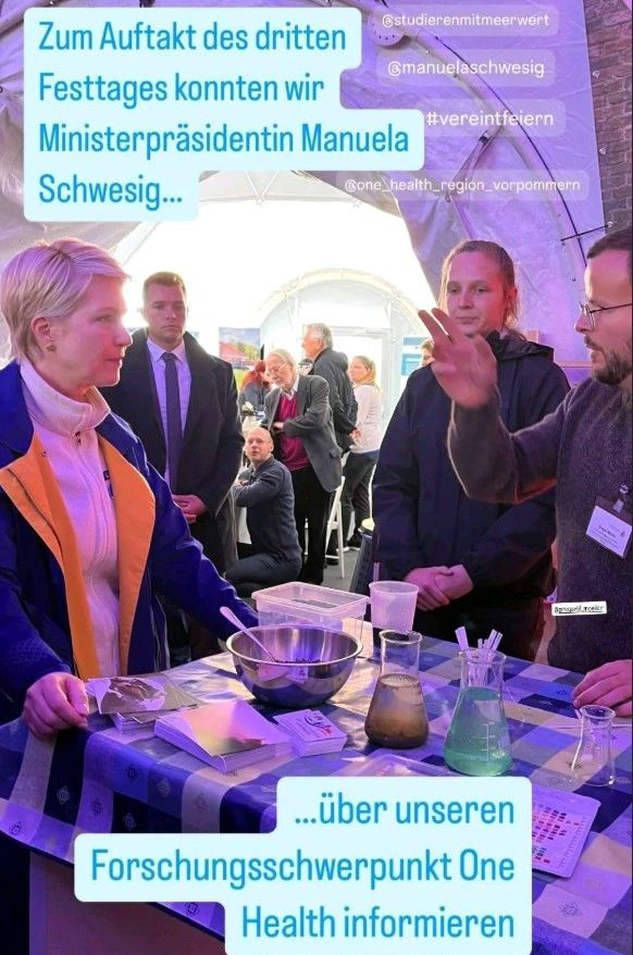Mehrere Personen stehen an einem Mitmachstand der One-Health-Region Vorpommern beim Bürgerfest zum Tag der Deutschen Einheit in Schwerin. Auf dem Tisch liegen Laborutensilien und Materialien zur anschaulichen Erklärung des One-Health-Ansatzes, während ein Gespräch stattfindet.
