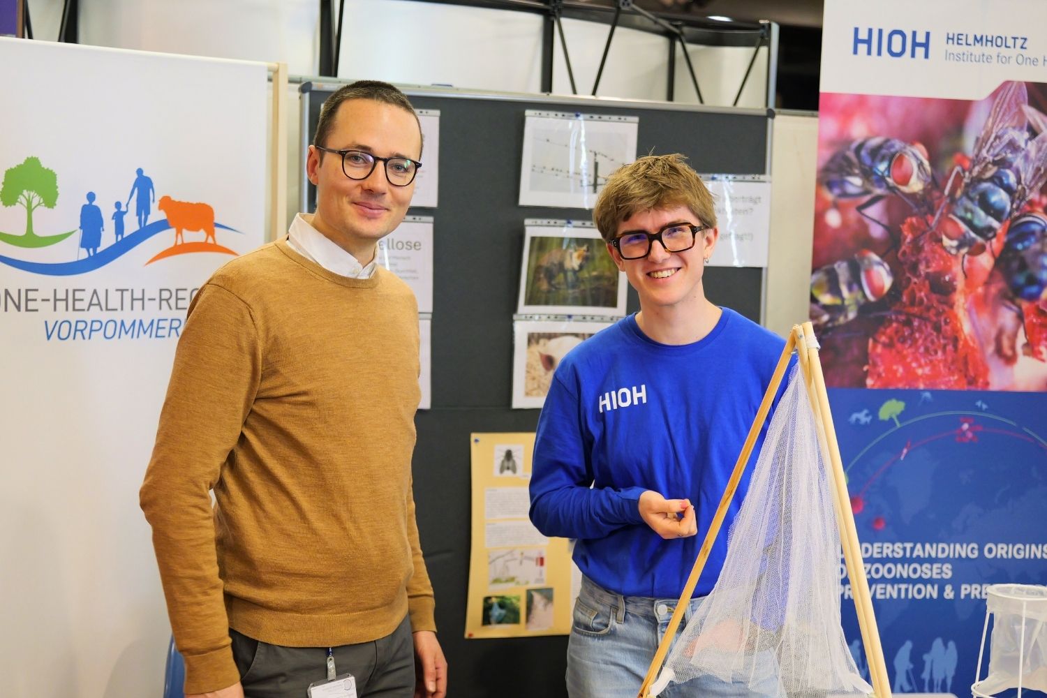 Zwei Personen stehen am Infostand der One Health Region Vorpommern auf der Gesundheitsmesse in Torgelow. Eine Person hält ein Fangnetz für Fliegen in der Hand. Im Hintergrund sind Informationsposter zum One-Health-Ansatz und ein Roll-up des Helmholtz Institute for One Health (HIOH) zu sehen.