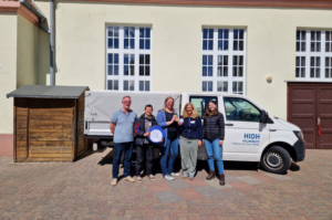 Fünf Personen stehen vor einem Fahrzeug des Helmholtz-Instituts für One Health auf dem Schulhof der Pestalozzi-Grundschule Torgelow. Eine Person hält ein rundes Schild mit der Aufschrift „Eine Gesundheit“. Im Hintergrund ist das Schulgebäude zu sehen.
