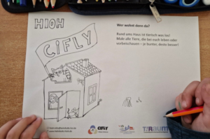 Kind hält Buntstifte über einer Ausmalvorlage des Projekts „CiFly“. Die Vorlage zeigt ein Haus mit verschiedenen Tieren und die Frage „Wer wohnt denn da?“. Auf dem Tisch liegen Stifte und Federmappen.