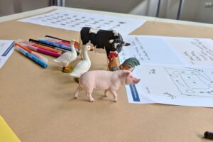 Auf einem Arbeitstisch stehen Tierfiguren von Schwein, Kuh, Gänsen und Hahn zwischen bunten Stiften und handschriftlich beschrifteten Workshop-Blättern mit Skizzen.