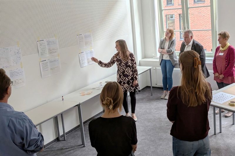 Teilnehmende einer Kreativwerkstatt stehen vor einer Wand mit aufgehängten Arbeitsblättern und besprechen gemeinsam die Ergebnisse.