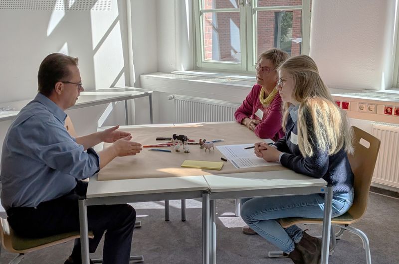 Drei Teilnehmende sitzen während der Kreativwerkstatt an einem Tisch und diskutieren miteinander. Auf dem Tisch liegen Stifte, Arbeitsblätter und kleine Tierfiguren.