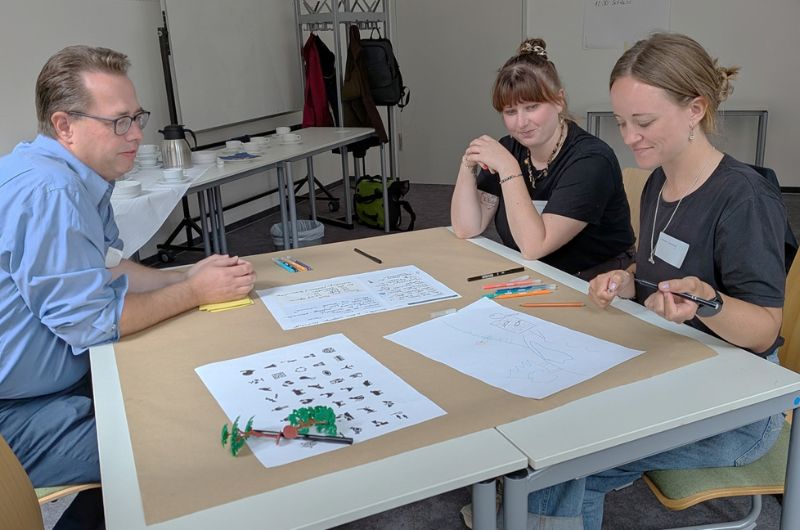 Drei Teilnehmende arbeiten gemeinsam an einem Tisch mit Skizzen, Stiften und Tierfiguren und entwickeln Ideen für die Kreativwerkstatt.