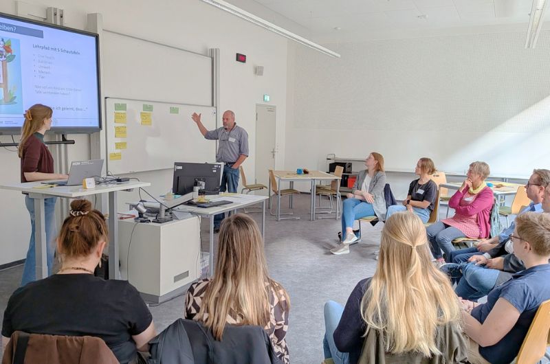 Teilnehmende sitzen im Plenum einer Kreativwerkstatt. Ein Teilnehmer spricht vor der Gruppe, während an Bildschirm und Pinnwand Arbeitsergebnisse präsentiert werden.