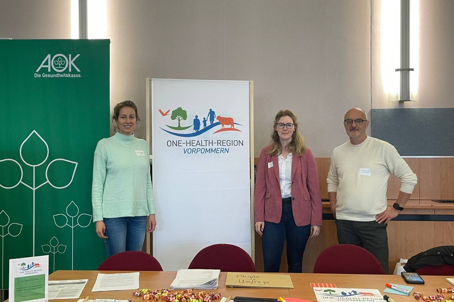 Drei Vertreterinnen und Vertreter der One-Health-Region Vorpommern stehen am Informationsstand beim Markt der Möglichkeiten der 4. Kommunalen Gesundheitskonferenz in Grimmen. Im Hintergrund sind ein Roll-up der One-Health-Region Vorpommern sowie ein Banner der AOK zu sehen.