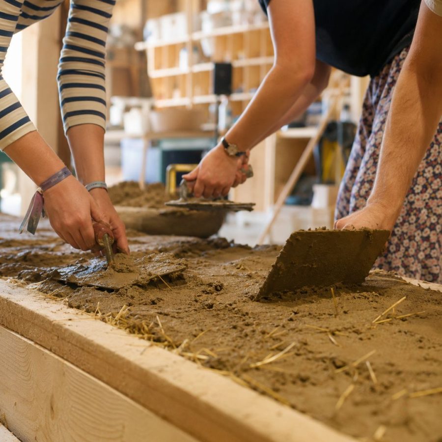 Drei Personen verarbeiten gemeinsam Lehm und Stroh auf einer großen Arbeitsfläche mit Maurerkellen.