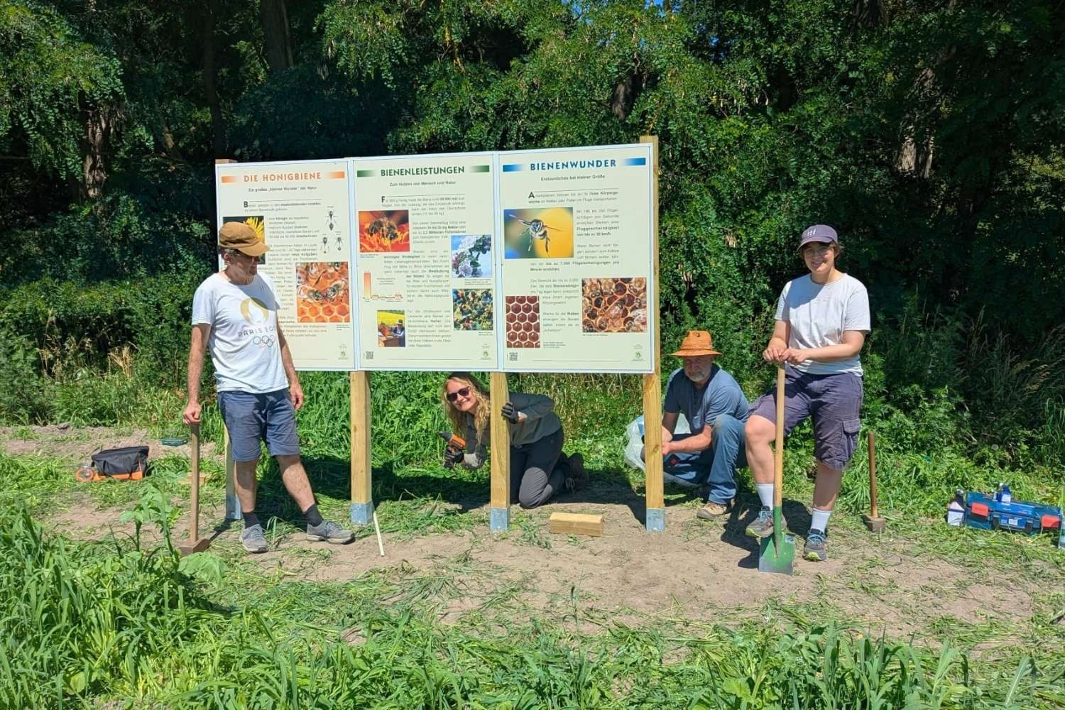 Vier Personen stehen vor drei neu aufgebauten Informationstafeln eines Bienen-Lehrpfads am Waldrand. Zwei halten Spaten, zwei befestigen die Tafeln. Die Schilder informieren über Honigbienen, ihre Leistungen für Mensch und Natur sowie besondere Eigenschaften von Bienen. Werkzeug und Baumaterial liegen auf dem Boden – die Tafeln wurden gerade aufgebaut.