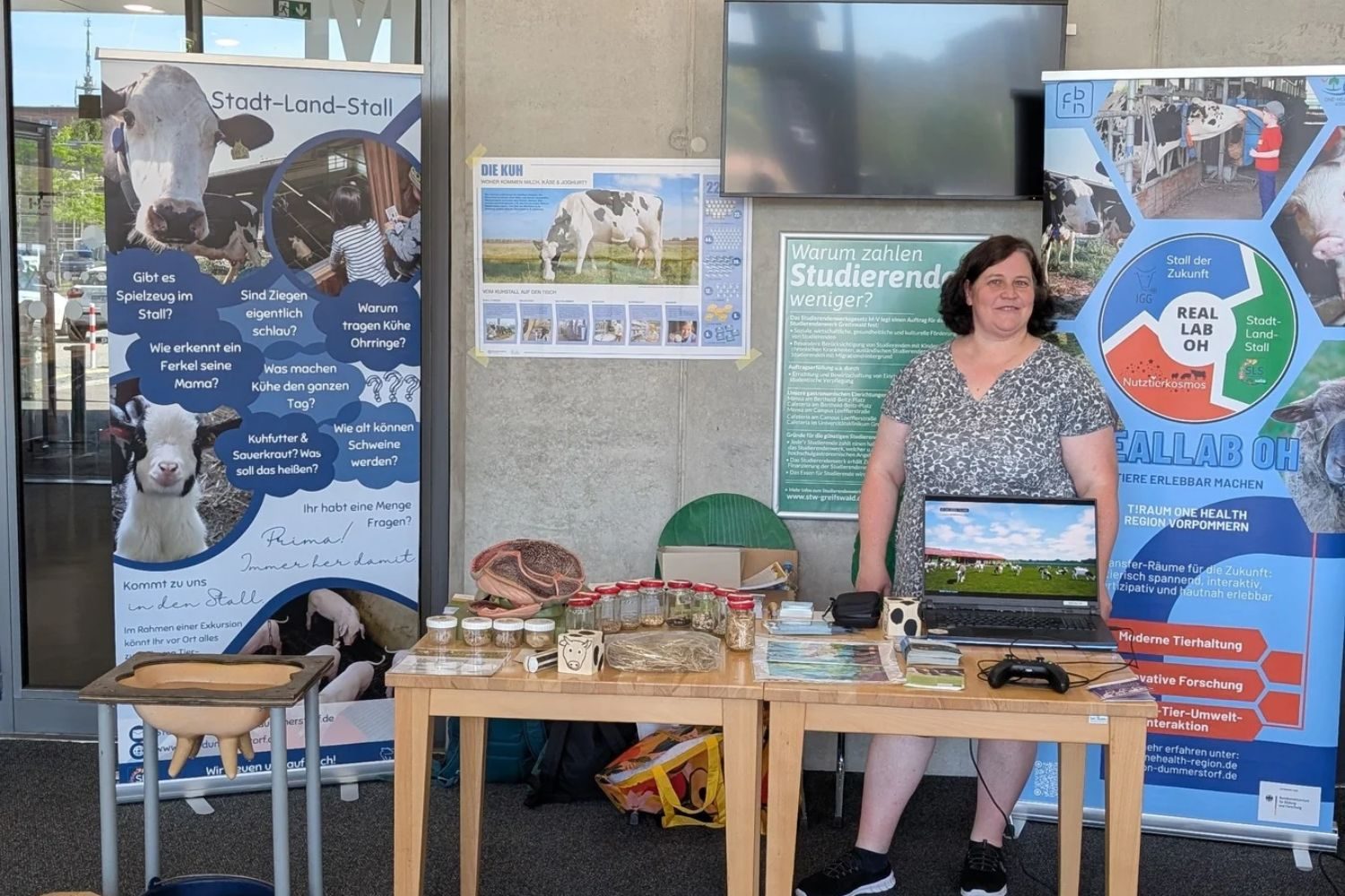 Eine Mitarbeiterin des Projekts Reallab OH am Informationsstand beim Tag der Wissenschaft 2026 in Greifswald. Auf den Tischen sind ein Eutermodell, ein Pansenmodell, Futterproben und Informationsmaterialien ausgestellt. Ein Laptop zeigt einen virtuellen Kuhstall.
